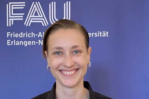 Paulina Jo Pesch vor einem blauen Banner mit dem Logo der FAU, lächelnd, am Tag ihrer Ernennung zur Juniorprofessorin.